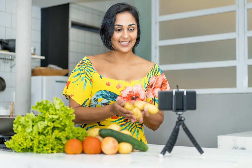 Mulher jovem com traços indianos de cabelos curtos e lisos, usando uma blusa amarela e segurando algumas batatas. Ela sorri para um celular que está filmando, apoiado em um tripé em uma cozinha.