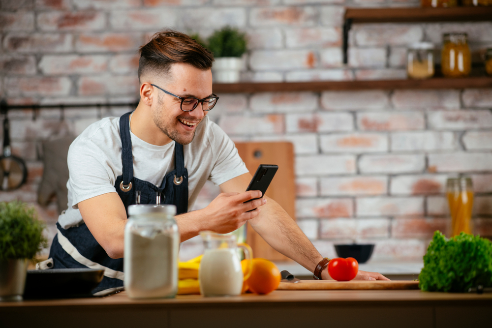 Fundo desfocado de uma cozinha com paredes de tijolinhos. Em foco, um homem branco e jovem de cabelos curtos e lisos, usando óculos. Ele usa uma camisa branca e, por cima, um avental preto. Ele está sorrindo enquanto olha para a tela de um celular em sua mão direita.