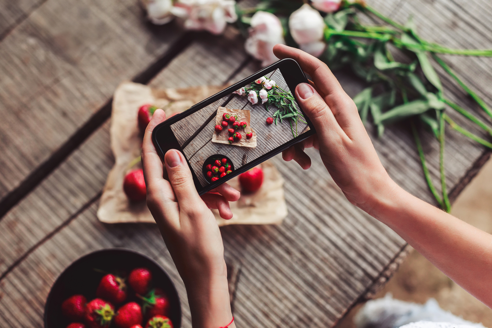 Fundo desfocado. Em foco, duas mãos de uma pessoa branca segurando um celular e fazendo uma foto de alguns tomates que estão por cima de um papel pardo, em uma mesa.