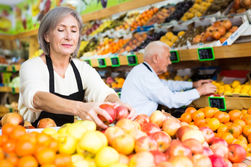 um homem e uma mulher, ambos idosos, organizando frutas na prateleira de um hortifruti