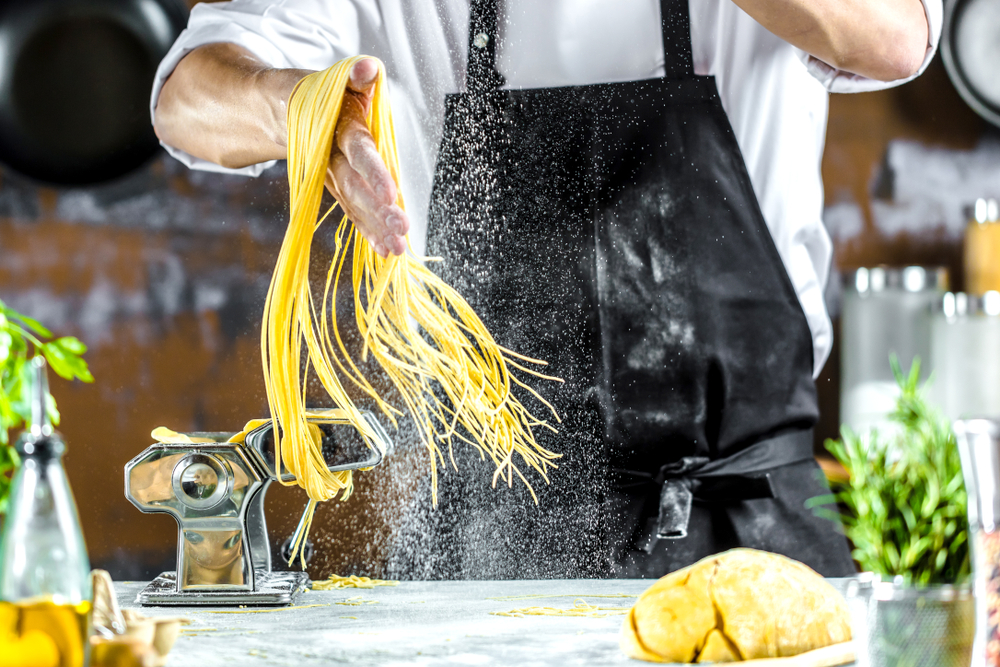 Chef usando um avental preto, fazendo macarrão espaguete com uma máquina de macarrão na mesa da cozinha com alguns ingredientes ao redor.