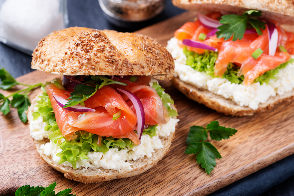 Imagem desfocada com dois sanduíches de salmão defumado com cream cheese: um sanduíche está na frente do outro, e os dois estão em cima de uma bandeja de madeira.